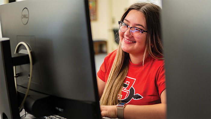 An SDSU student, wearing a t-shirt with the school logo, looks at a computer screen.